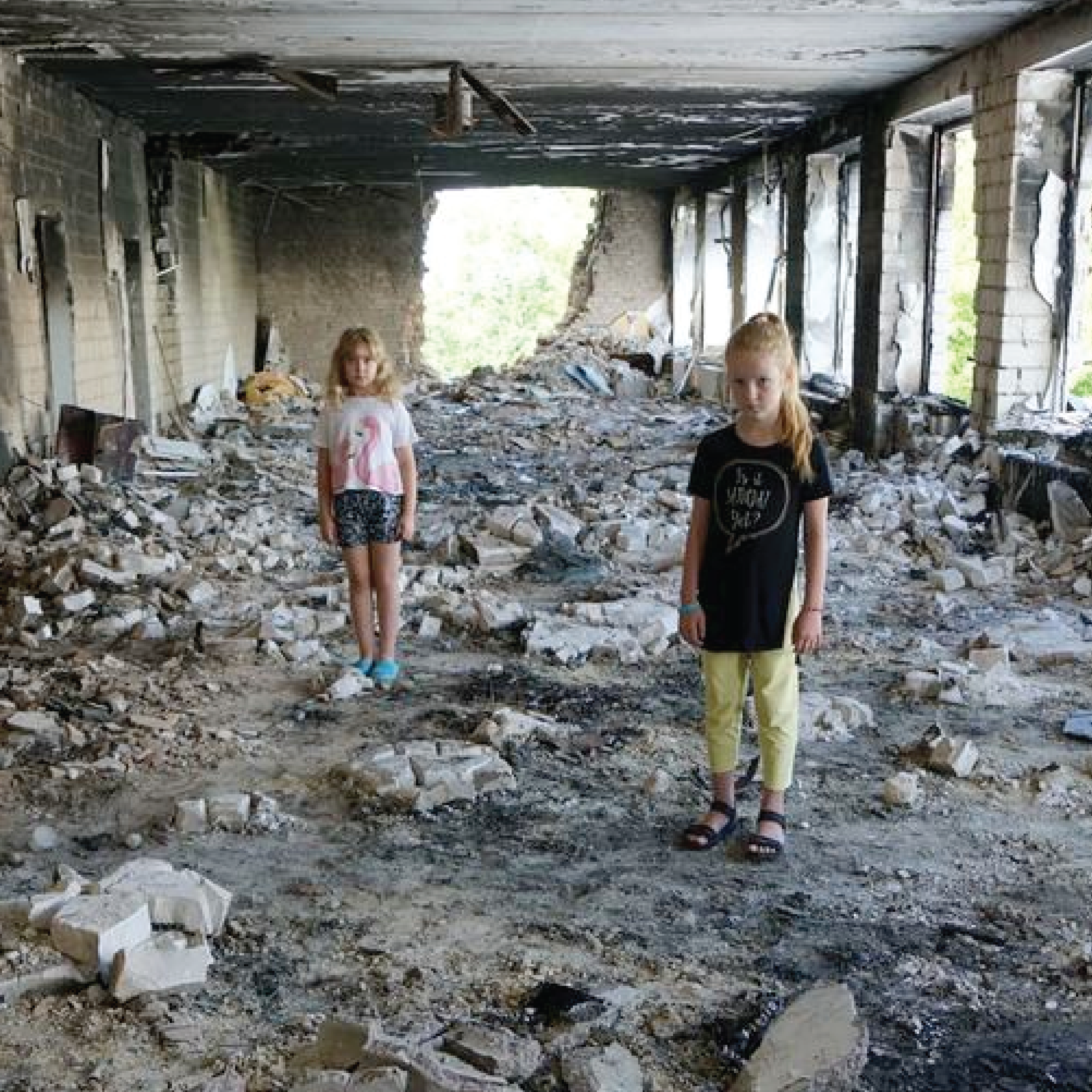 Eight-year-old friends stand amidst rubble in their damaged school in Buzova, Ukraine.